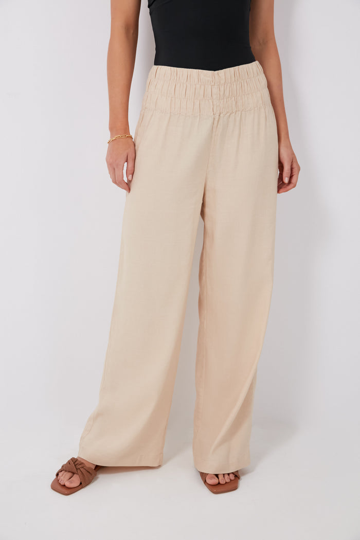 A person confidently stands in onia high-waisted, wide-leg Sand Air Smocked Pants (linen blend), paired with a black sleeveless top, brown sandals, and a gold bracelet.