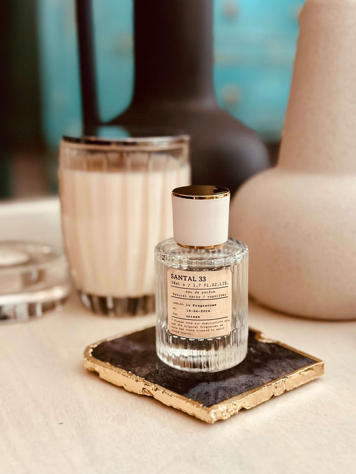 Sandalwood & Cardamom Eau de Parfum by LU Fragrances — 50ml glass bottle lying on its side on a gold-edged black stone coaster, with a white candle and ceramic vases in soft focus behind