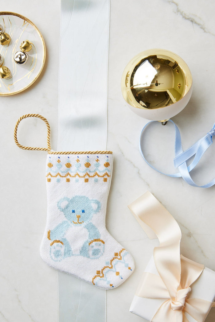 A small hand-stitched Bauble Stockings Christmas stocking with a blue teddy bear design is displayed on marble with a gold ornament, branded ribbon, bells, and a gift box.