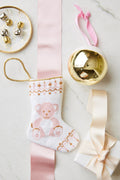 A small white hand-stitched Christmas stocking by Bauble Stockings featuring a teddy bear, a gold ornament, jingle bells on a dish, and a ribbon-wrapped gift are arranged on a marble surface.
