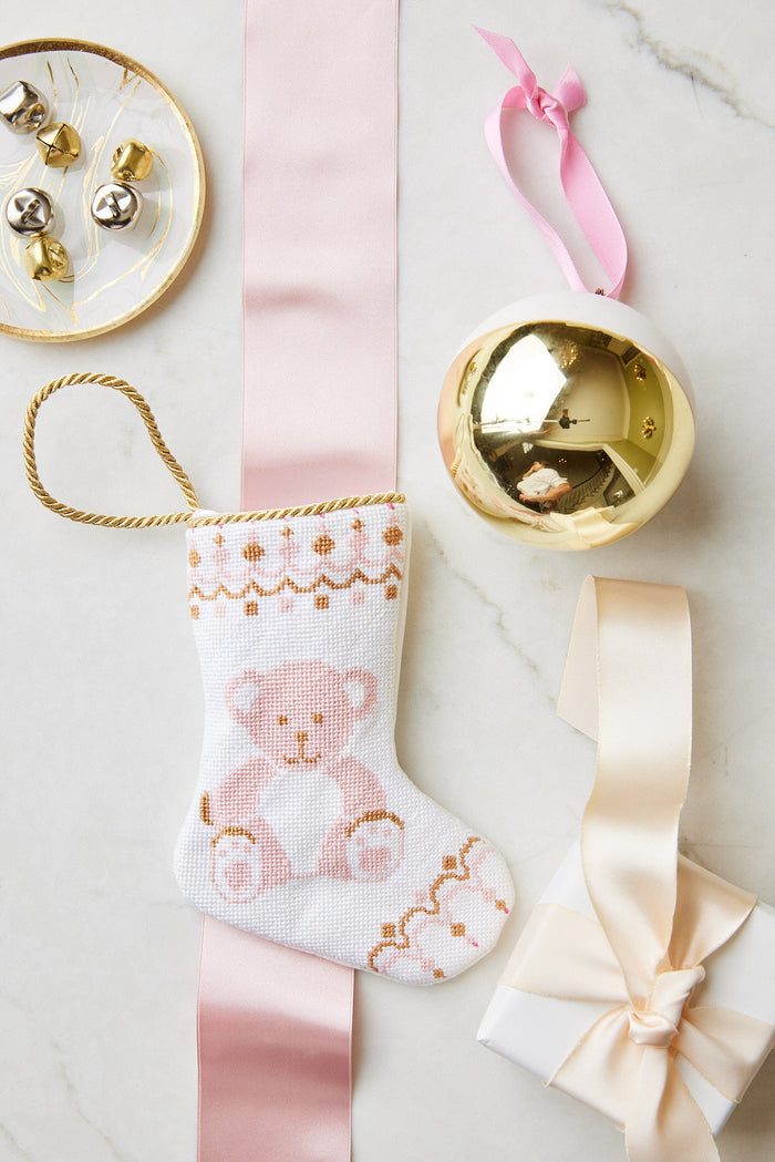A small white hand-stitched Christmas stocking by Bauble Stockings featuring a teddy bear, a gold ornament, jingle bells on a dish, and a ribbon-wrapped gift are arranged on a marble surface.