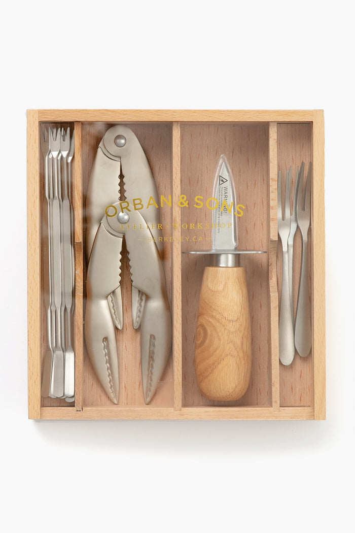 The Orban and Sons wooden box contains seafood tools—a crab cracker, wooden-handled oyster knife, seafood forks, and cocktail forks—each organized in its own compartment.