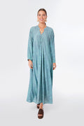 A woman wearing a blue patterned bohemian maxi dress with long sleeves by Natalie Martin, paired with brown sandals and statement earrings.
