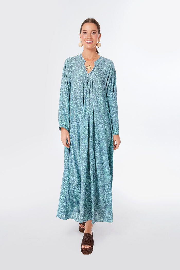 A woman wearing a blue patterned bohemian maxi dress with long sleeves by Natalie Martin, paired with brown sandals and statement earrings.