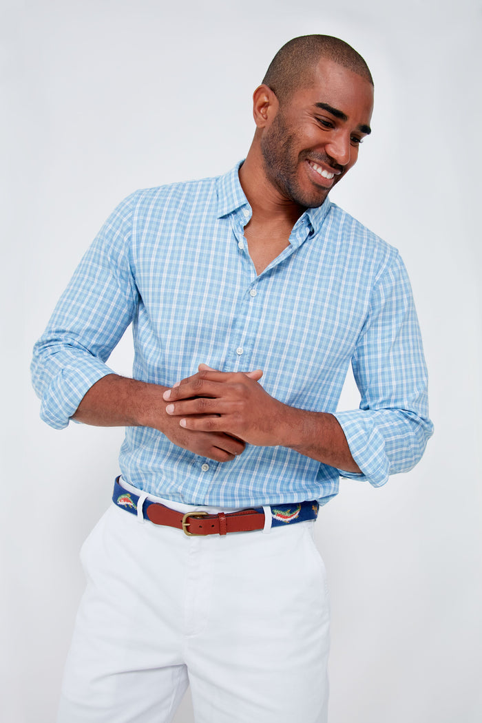 A man wearing a light blue checkered, wrinkle-resistant Supima cotton Faherty shirt and white pants stands smiling with his hands clasped together.