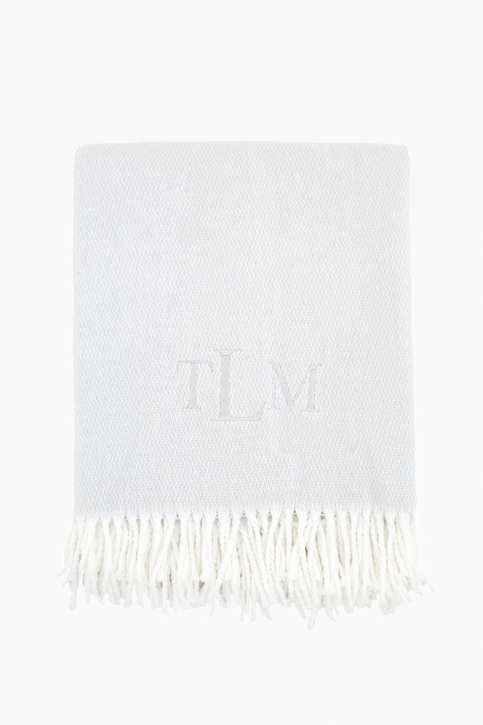 A light grey Matouk Silver Pezzo Throw features a white fringe trim and the monogram TLM embroidered at the center.