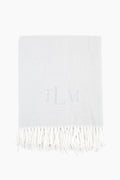 A light grey Matouk Silver Pezzo Throw features a white fringe trim and the monogram TLM embroidered at the center.