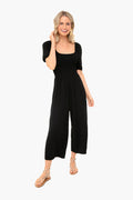 A woman stands smiling, wearing a BB Dakota black short-sleeve smocked jumpsuit with wide-leg pants and tan sandals.