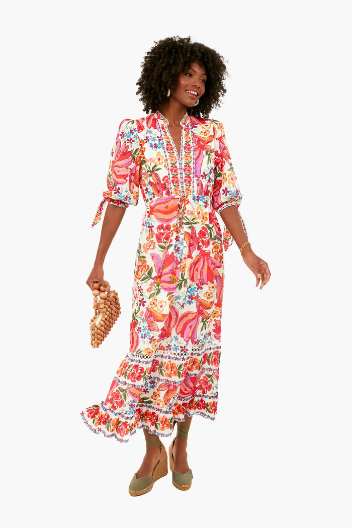 A woman in a Farm Rio tropical print maxi dress with puff sleeves holds a beaded handbag and stands in wedge shoes.