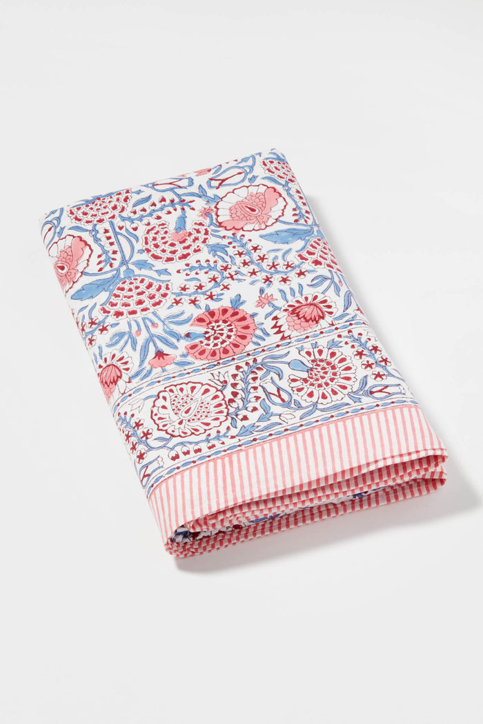 A folded Locust Walk Sunset Poppy Vines cotton tablecloth, featuring a red and blue floral-and-leaf pattern and pink and white striped border, is neatly displayed.