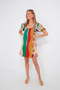A woman wears a vibrant Farm Rio mini dress with short sleeves and abstract floral patterns, made from organic cotton, styled with beige sandals and pink earrings.