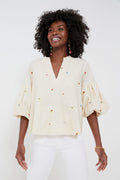 A person wearing a cream-colored, puff-sleeve embroidered blouse with colorful details from Hyacinth House, paired with white pants, stands smiling.