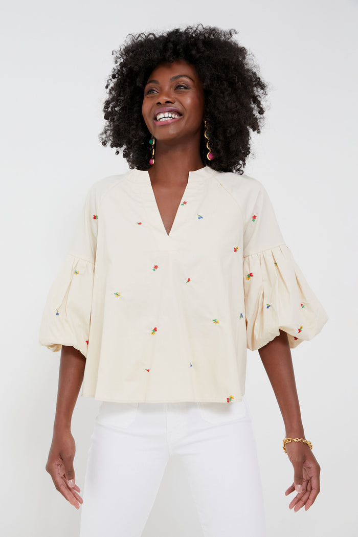 A person wearing a cream-colored, puff-sleeve embroidered blouse with colorful details from Hyacinth House, paired with white pants, stands smiling.