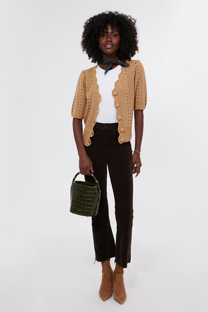A woman models Hyacinth House women's fashion in a tan crochet cardigan with short sleeves, white top, dark brown pants, tan ankle boots, black neck scarf, and green handbag.