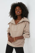 A woman with curly hair wears a beige Varley Half Zip sweater with a wide collar, black pants, and gold jewelry, showcasing a chic performance-fashion style.