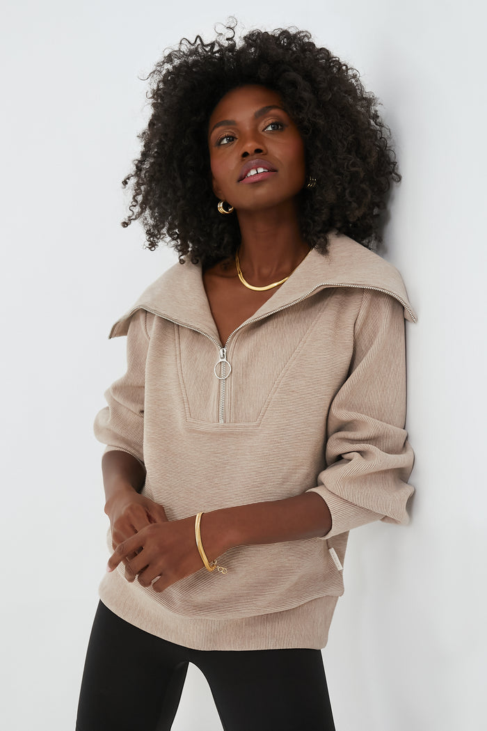 A woman with curly hair wears a beige Varley Half Zip sweater with a wide collar, black pants, and gold jewelry, showcasing a chic performance-fashion style.