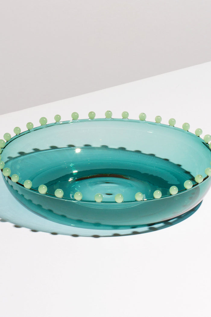 A shallow, translucent blue-green FAZEEK bowl made of hand-blown borosilicate glass with small round beads around the rim rests on a white surface.