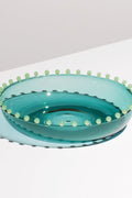 A shallow, translucent blue-green FAZEEK bowl made of hand-blown borosilicate glass with small round beads around the rim rests on a white surface.
