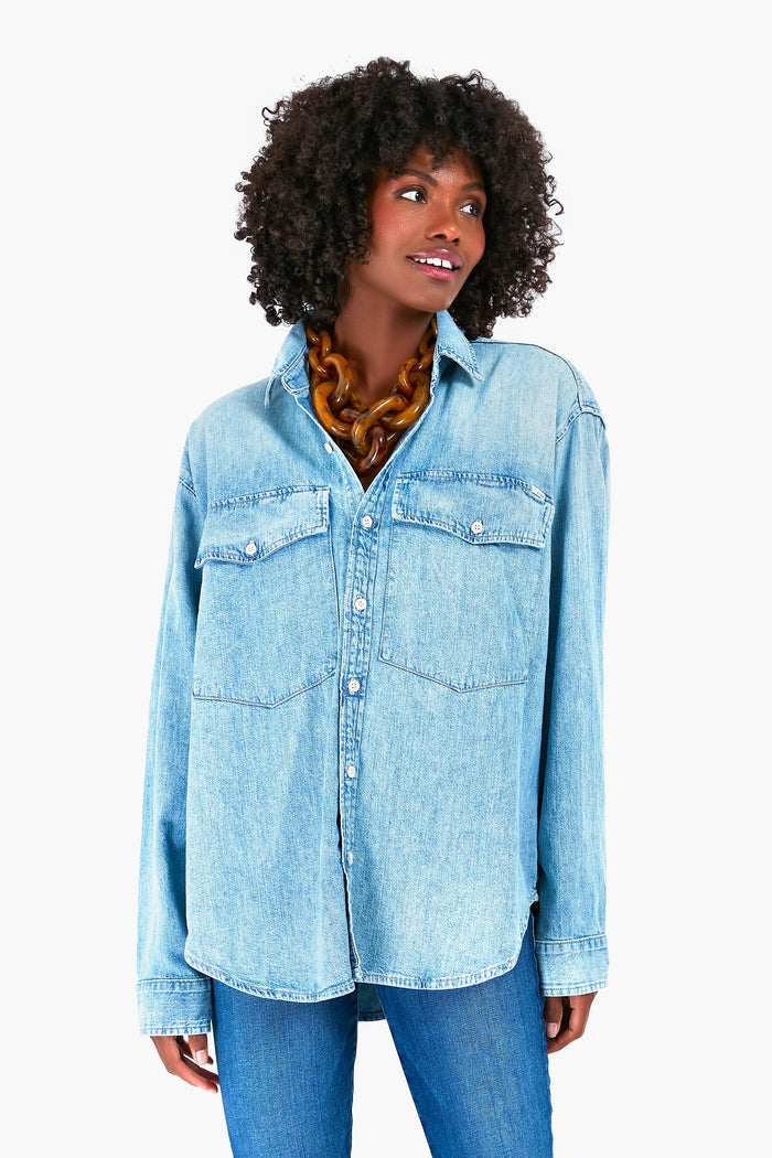 A woman wearing a light blue Double Flap Roomie Foxy Shirt and MOTHER jeans stands slightly to the side, sporting a chunky brown necklace and curly hair.