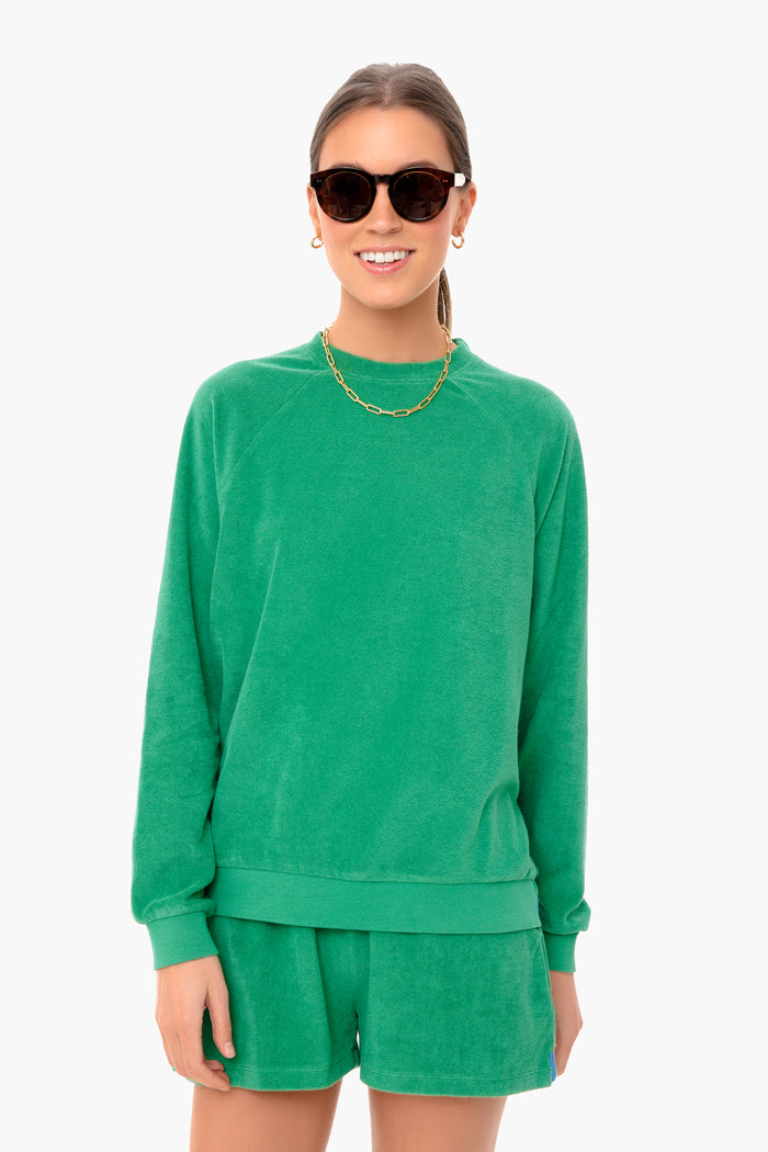 A woman wears a green oversized KULE long-sleeve sweatshirt with matching shorts, styled with dark sunglasses, gold earrings, and a gold chain necklace.