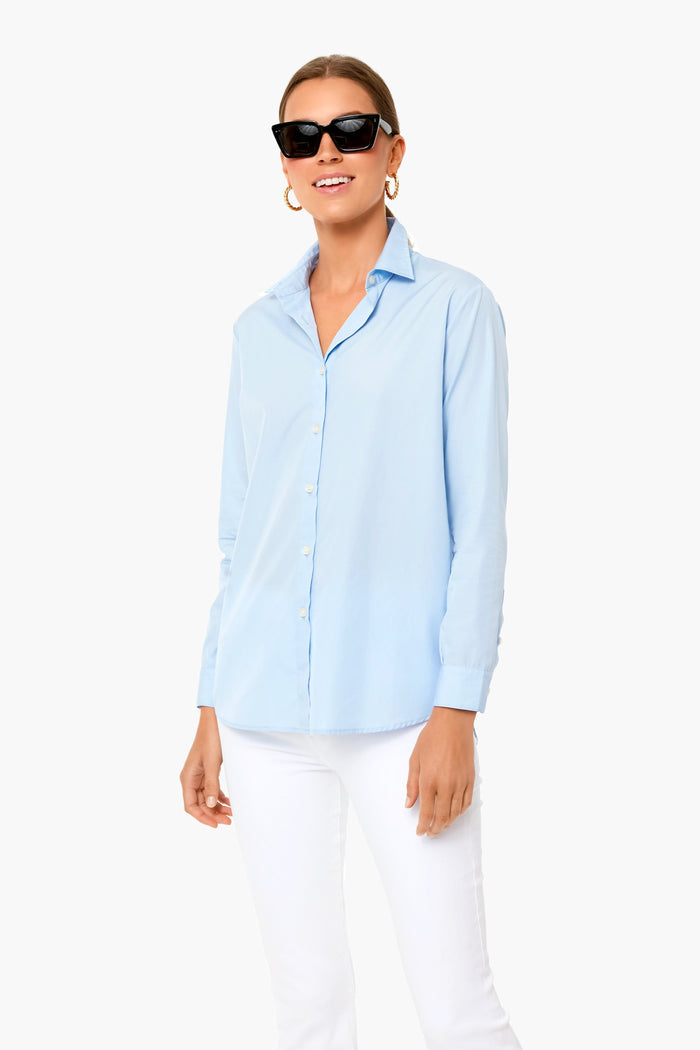 A woman in black sunglasses, gold hoop earrings, a KULE Italian cotton button-down shirt, and white pants stands confidently.