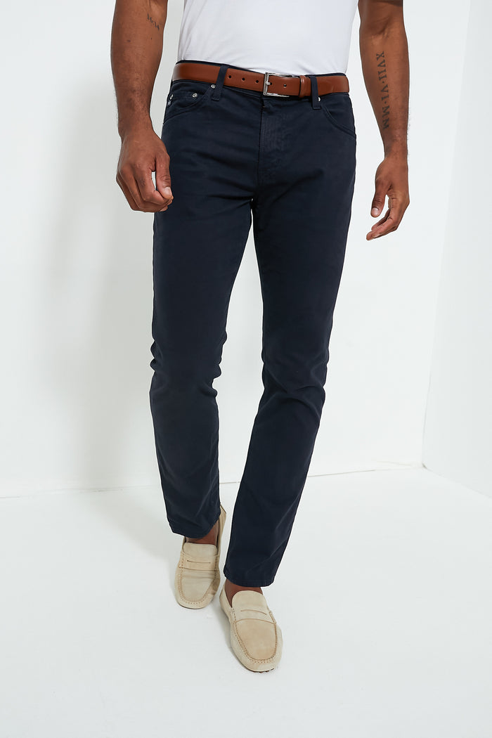A person wearing a white t-shirt, dark blue AG Jeans, a brown belt, and beige loafers.