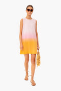 A woman in sunglasses wears a KULE ombré sunset dress in white, pink, and yellow, holding a straw bag and wearing flat sandals.