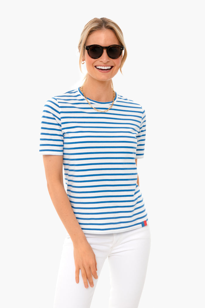 A woman smiles while wearing black sunglasses, gold necklaces, white pants, and a modern KULE white tee with royal blue stripes.