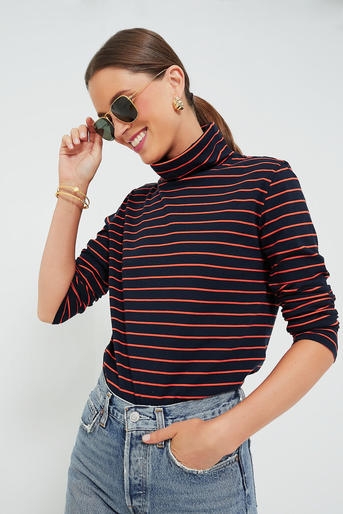 A person in a KULE navy and red striped turtleneck and jeans smiles while touching their sunglasses.