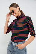 A person in a KULE navy and red striped turtleneck and jeans smiles while touching their sunglasses.