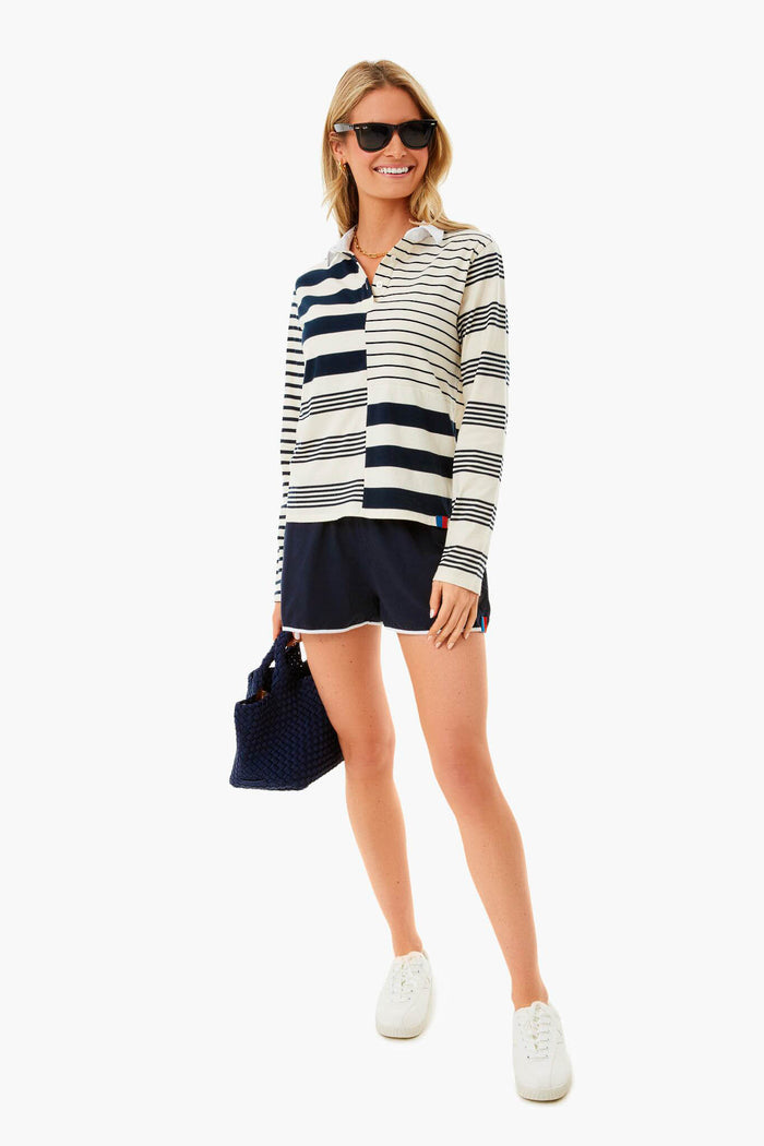 A woman wearing sunglasses, a KULE striped cotton rugby shirt, navy shorts, white sneakers, and holding a navy handbag stands and smiles.