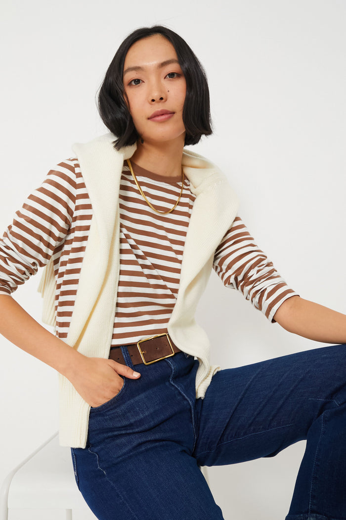 A person sits on a stool wearing a KULE brown and white striped tee, dark blue jeans, and a cream sweater draped over their shoulders.