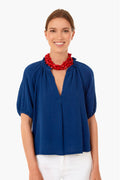 Wearing the XiRENA Tiara Blue Jules Top with a ruffled neckline and bubble sleeves, paired with white pants and a chunky red beaded necklace, she smiles confidently at the camera.