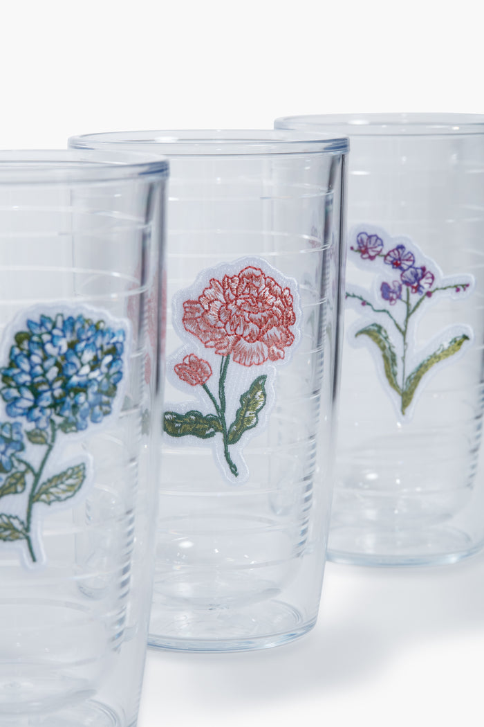 Three clear BPA-free Tervis Tumbler x Tuckernuck tumblers, each with an embroidered patch of a blue hydrangea, red carnation, or purple violet, arranged in a row.
