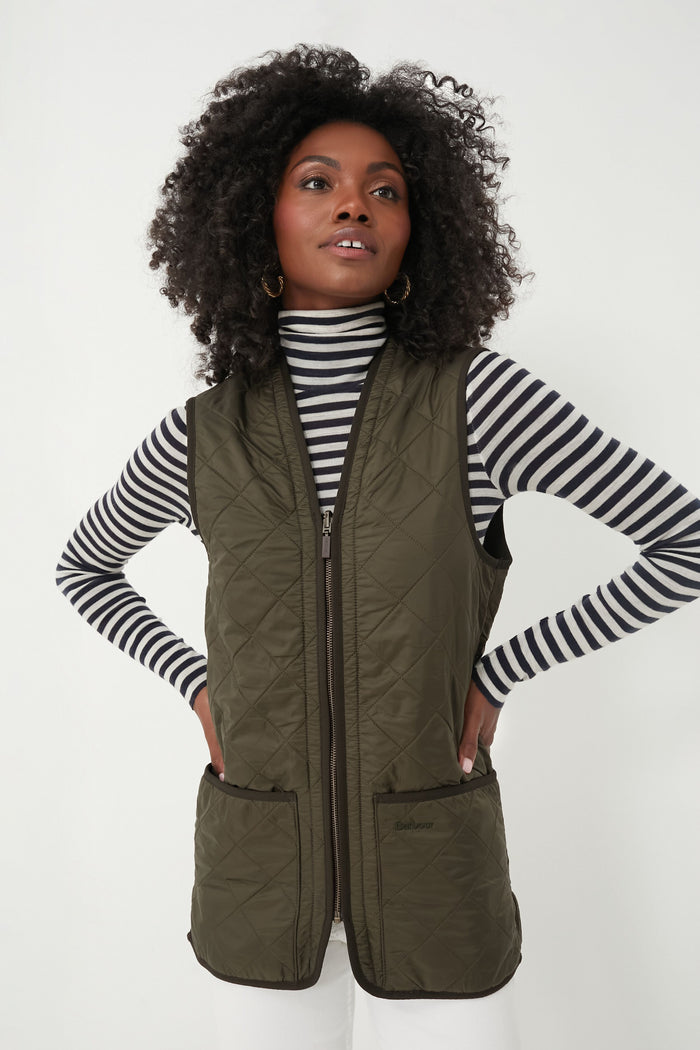 A woman stands with her hands on her hips, wearing a black-and-white striped turtleneck and a Barbour olive Polarquilt Waistcoat.