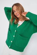 A woman wears a bright green Alex Mill Emma Cardigan featuring large front pockets and white buttons, paired with white pants.