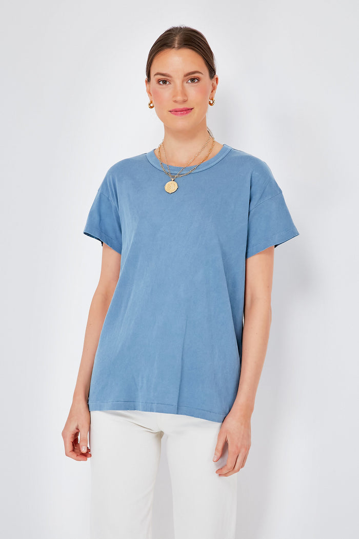 A woman wears THE GREAT. Vintage Cornflower Boxy Crew Tee in blue, styled with white pants, gold necklaces, and hoop earrings. This classic tee features a boxy fit and is made from 100% cotton for lasting comfort and style.