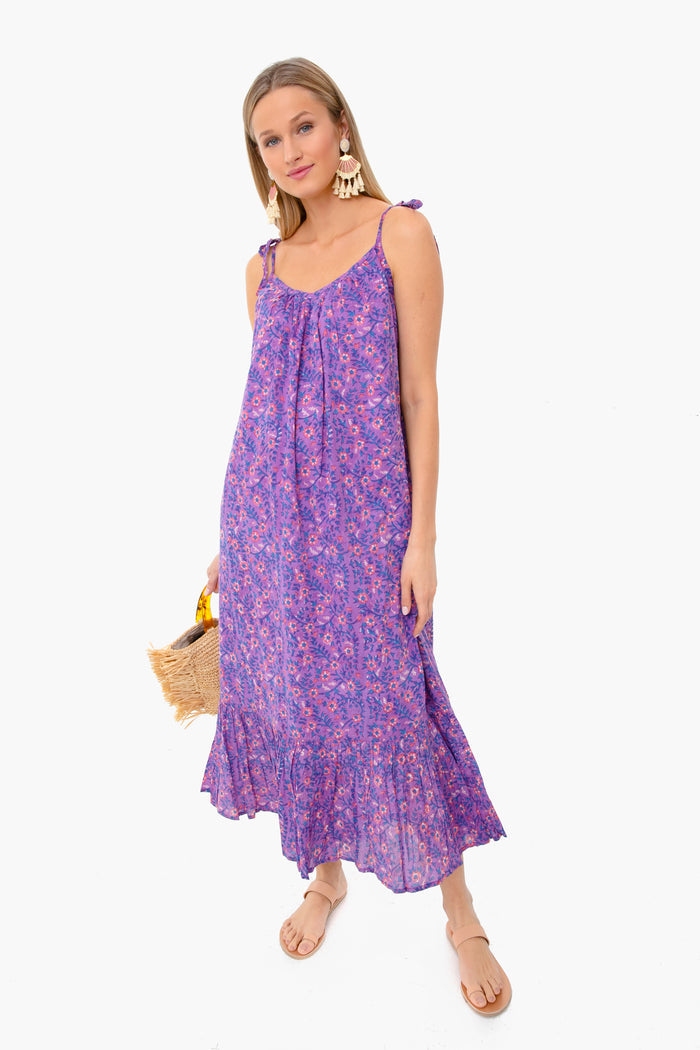 A woman wears a sleeveless purple floral India Sundress with shoulder ties from Love Tomas by Emerson Fry, styled with tan sandals, tassel earrings, and a straw clutch.