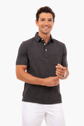 A man smiles in a dark Faherty organic cotton polo with a chest pocket and white pants, arms slightly bent. This classic men’s shirt blends comfort with sustainable apparel style.