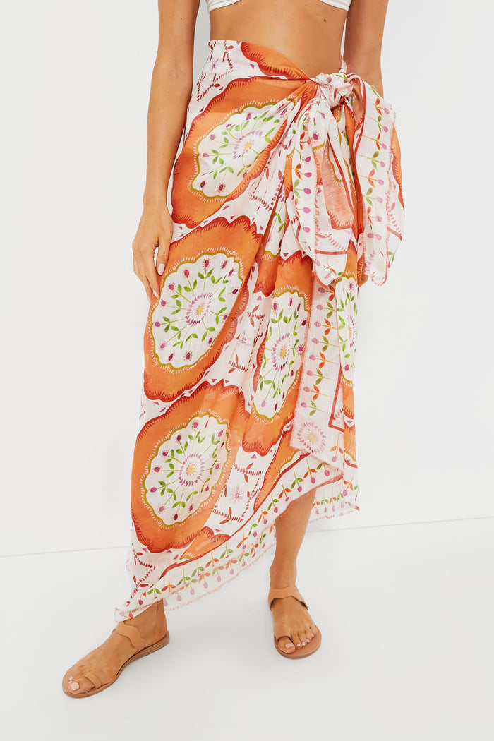 A person wearing an ASHA x Tuckernuck white and orange patterned wrap skirt with a large bow at the waist and tan sandals.