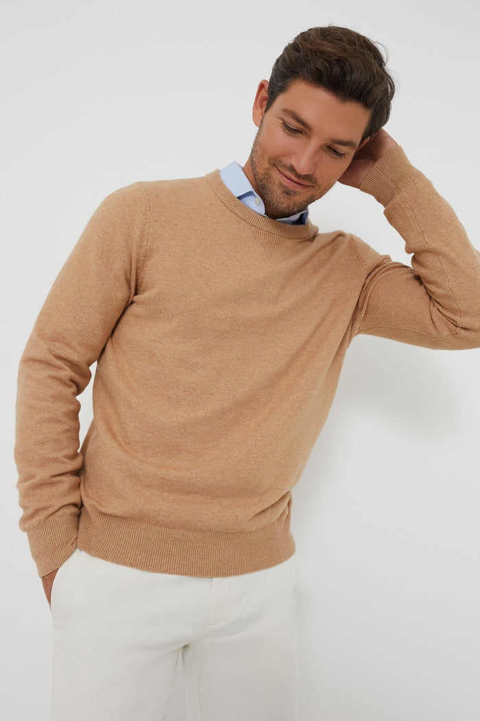 A man in a Faherty sustainable, moisture-wicking crewneck sweater layered over a blue dress shirt and white pants stands confidently, one hand resting on the back of his neck.