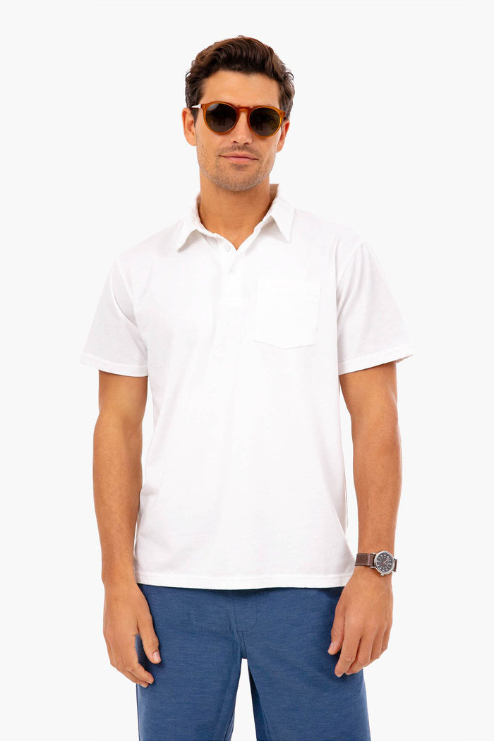 A man confidently poses wearing Fair Harbor sunglasses, a white recycled polyester shirt with a chest pocket, blue pants, and a wristwatch.