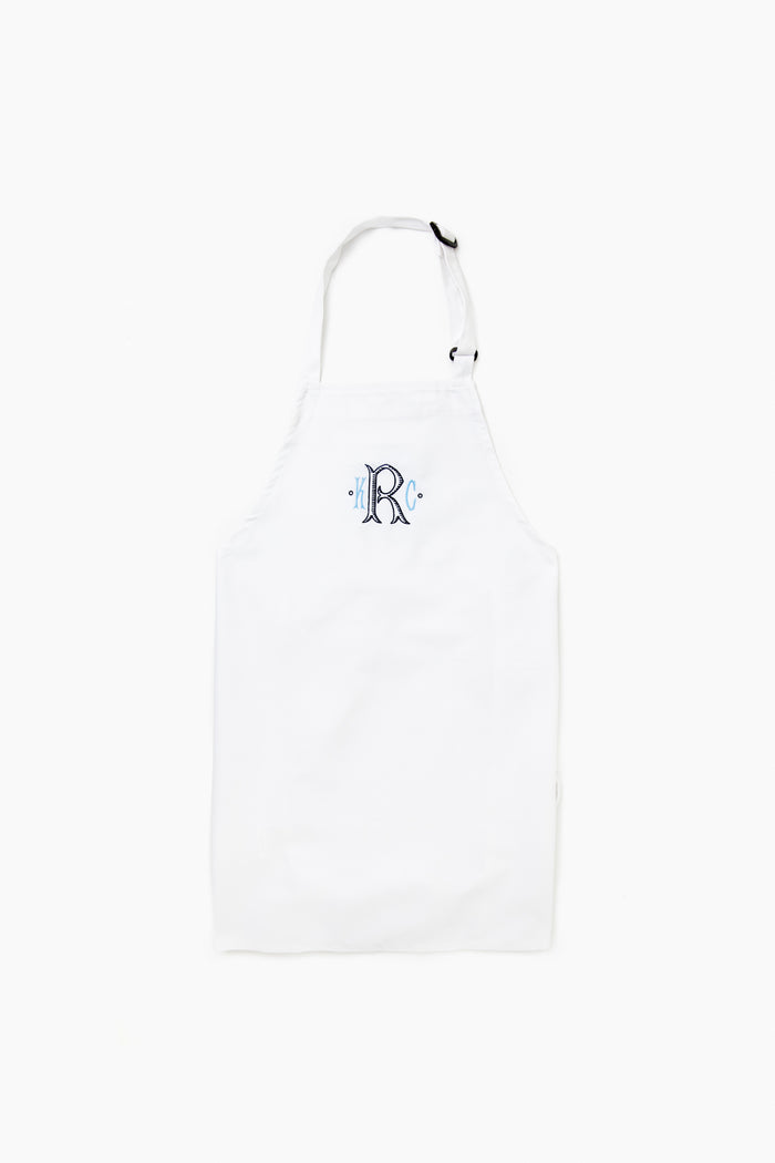 The Caton's Wharf white adjustable neck apron features a blue letter R monogram on the chest and is crafted from wrinkle-resistant fabric for durability and style.