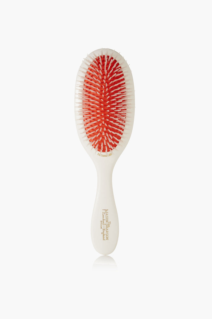 A Mason Pearson luxury hairbrush is shown, featuring a white handle, red cushioned base, and white bristles.