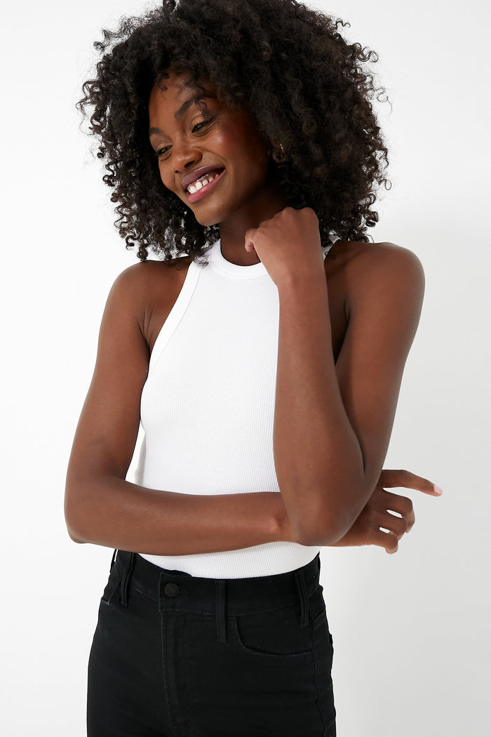 A woman with curly hair smiles confidently in an Anine Bing white organic cotton tank top and black jeans—effortlessly showcasing true wardrobe essentials.