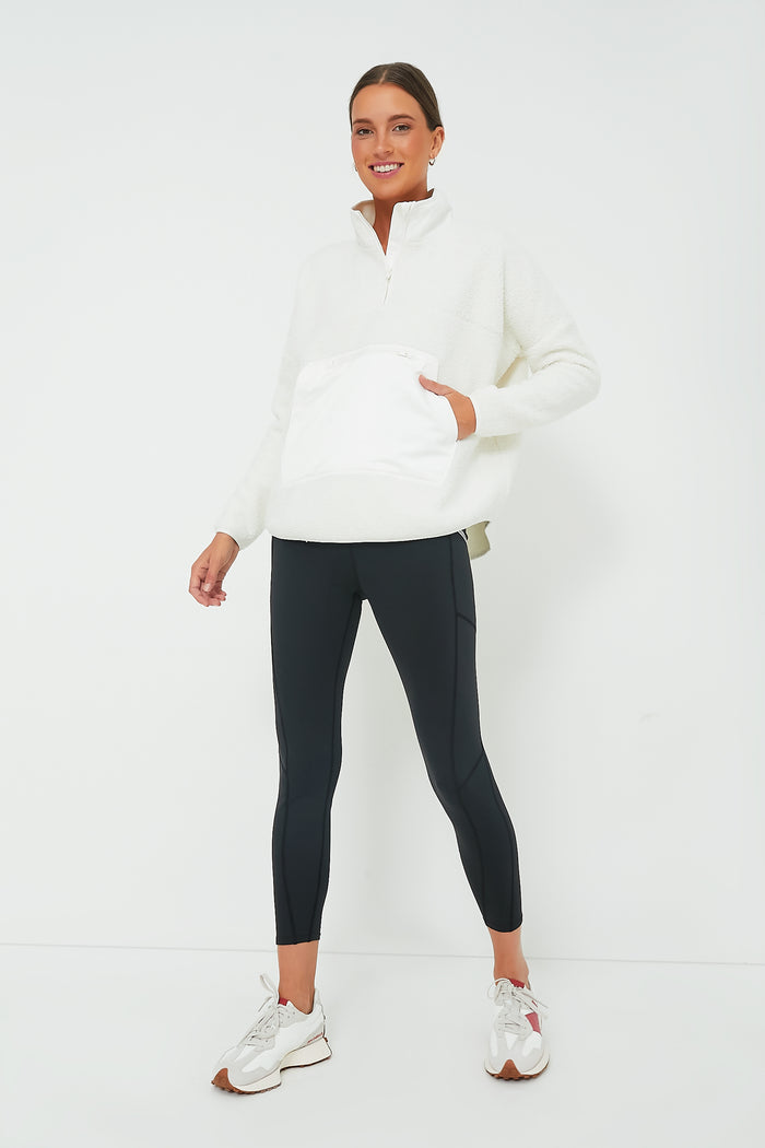 A woman stands against a white background wearing a Tnuck Sport White Fleece Cady Quarter Zip, black leggings, and white sneakers, with her hair pulled back and hands by her sides.