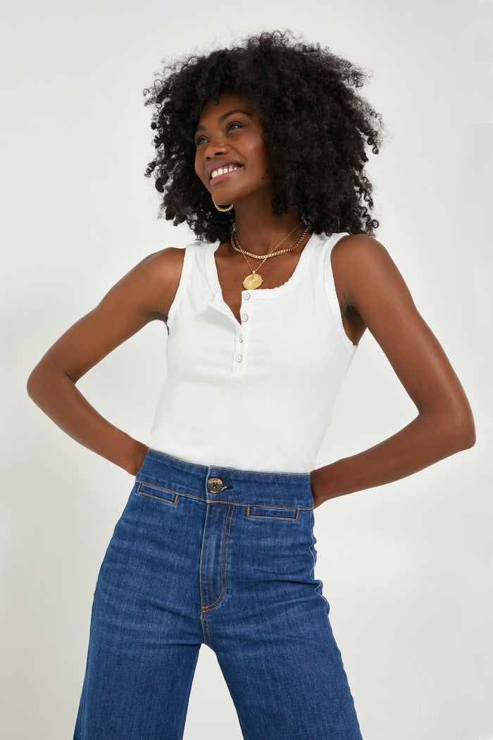 A woman stands with hands on hips, smiling in an ASKK NY white henley tank and timeless denim jeans, layered gold necklaces completing her look.