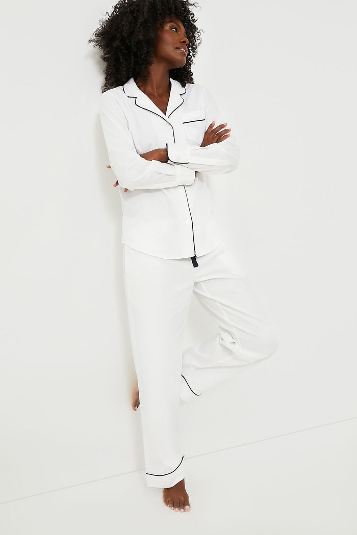 A person stands barefoot, arms crossed and one leg bent, wearing Petite Plume's white cotton pajama set with navy piping.