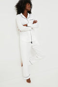 A person stands barefoot, arms crossed and one leg bent, wearing Petite Plume's white cotton pajama set with navy piping.