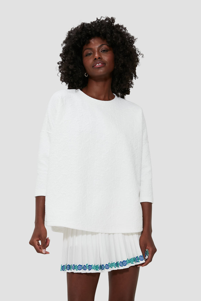 A woman wears a Tnuck Sport textured white quilted sweatshirt paired with a white pleated skirt featuring blue floral embroidery.